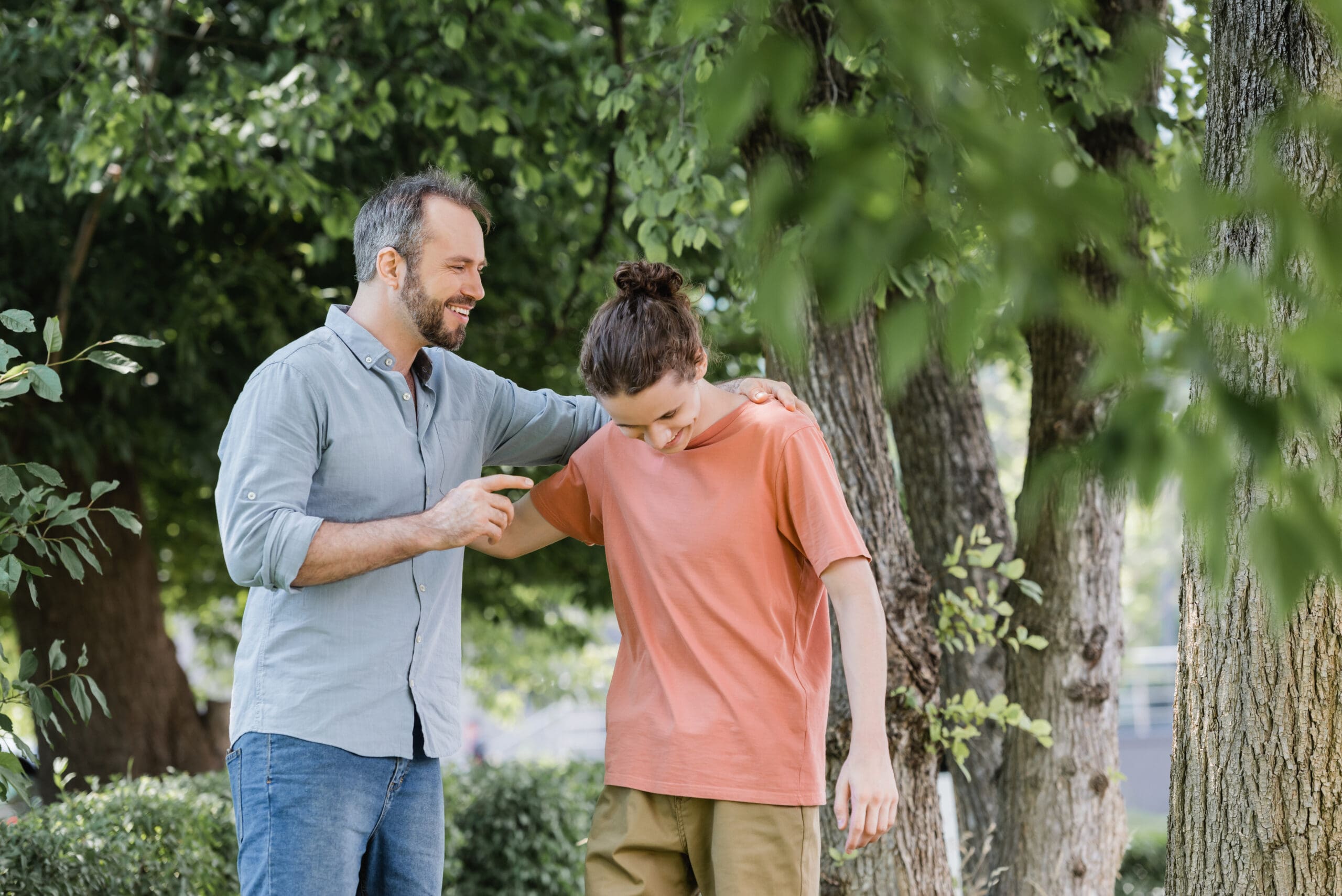 cheerful father with beard hugging happy teenager son in green park